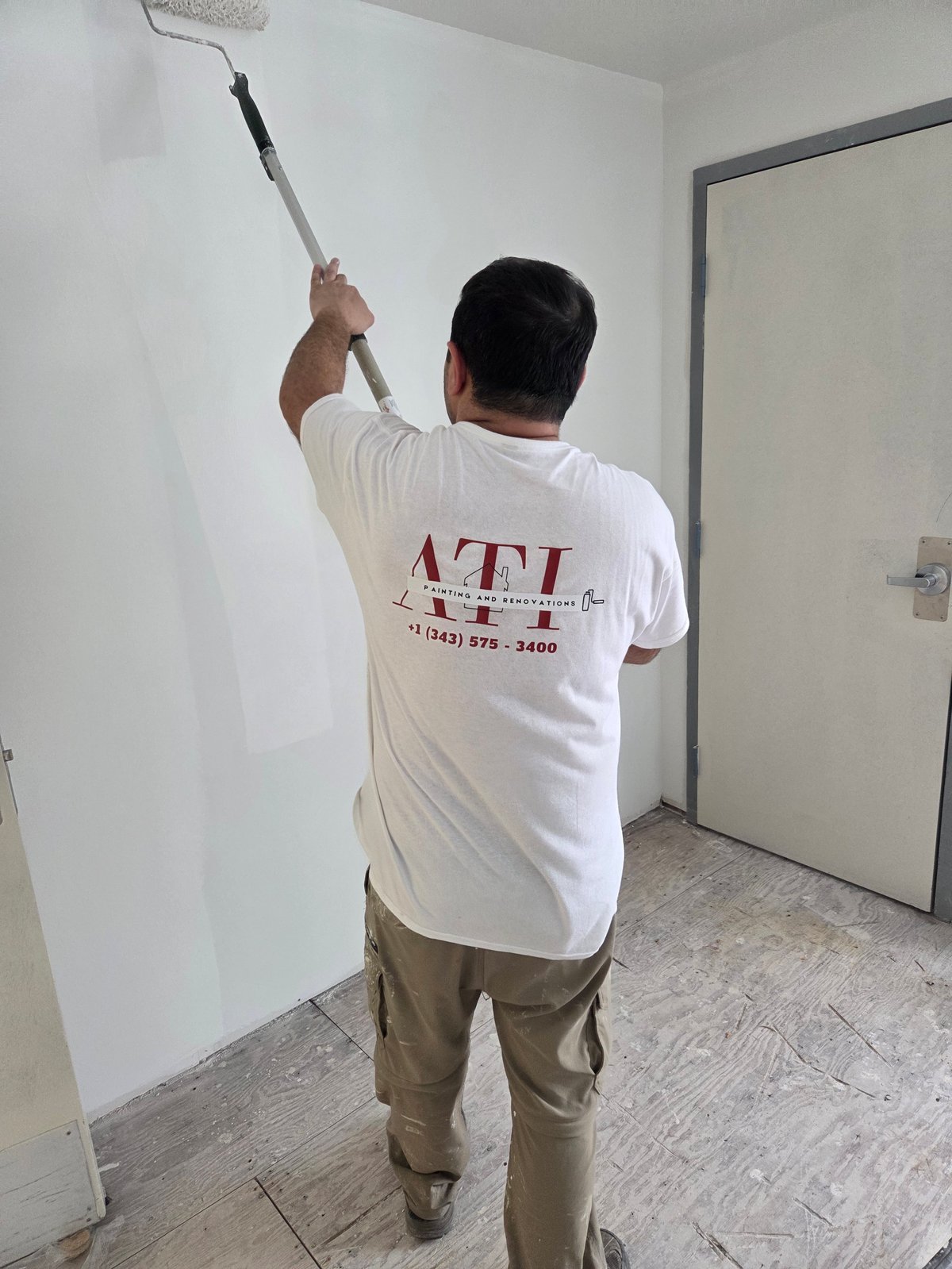 ATI crew member painting a wall with a roller in a bright interior room, seen from behind
